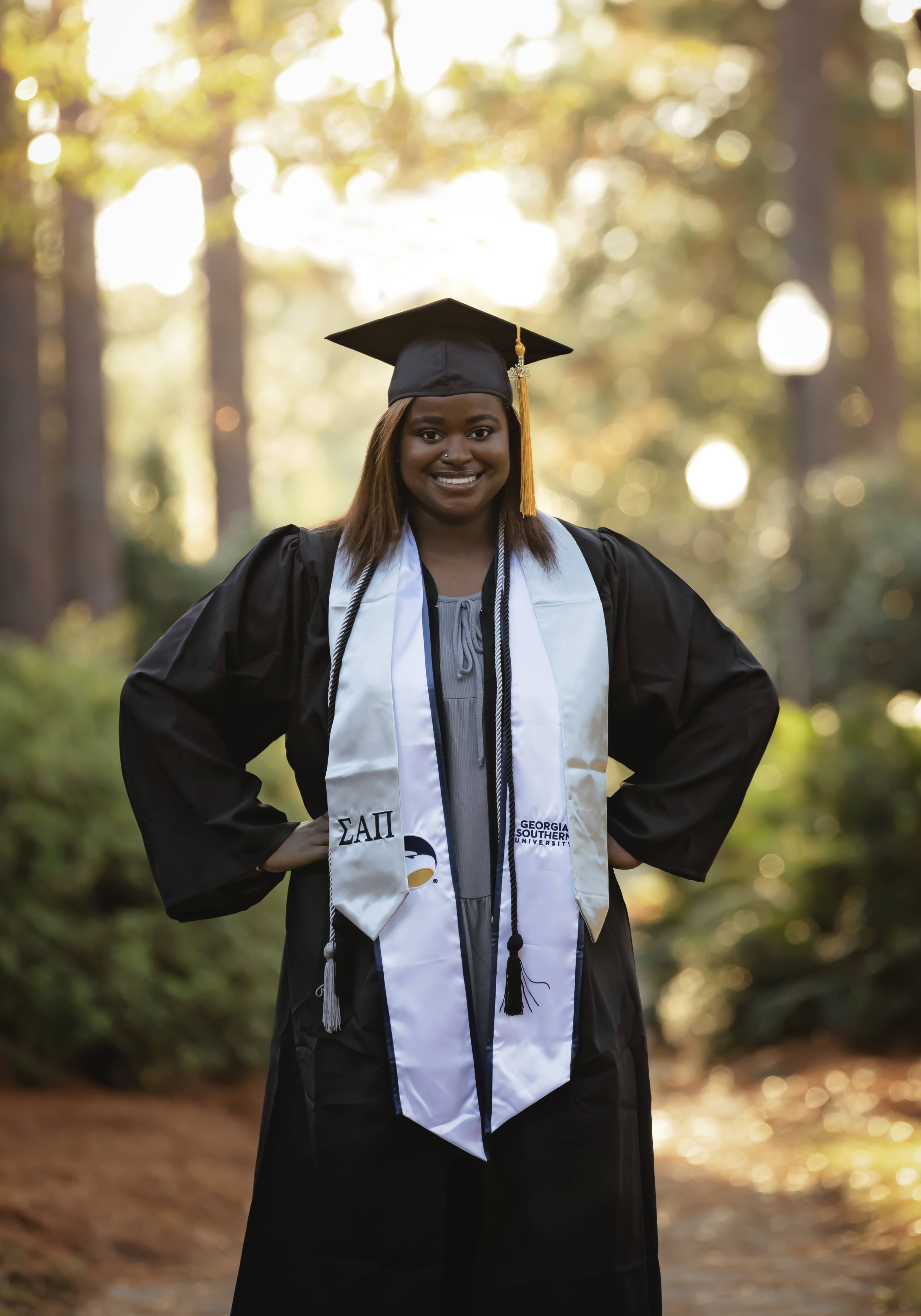 Graduate wearing graduation robes stands with trees in the background